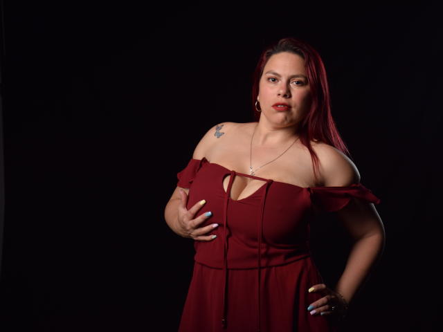 SalmaBrown with red hair, off-shoulder maroon dress, hand on her tits, tattoo on right shoulder, dark background, serious expression, necklace, light skin, medium build.