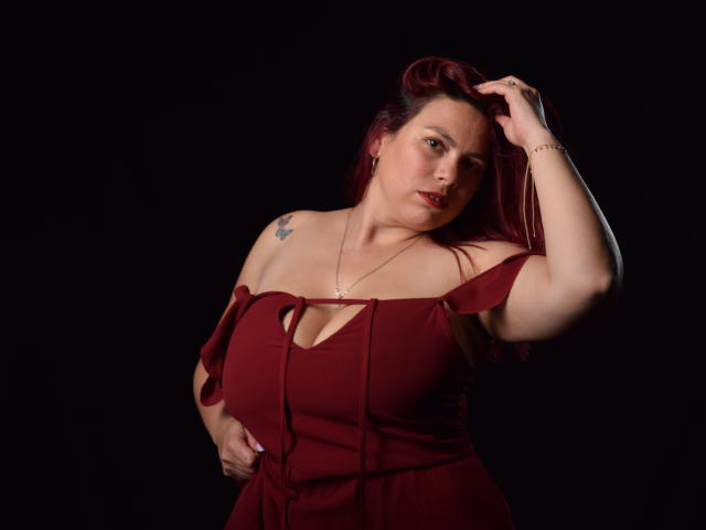 SalmaBrown with red hair, off-shoulder maroon dress, hand in hair, tattoo on left shoulder, necklace, red lipstick, black background, slightly overweight, confident, sexy, attractive.