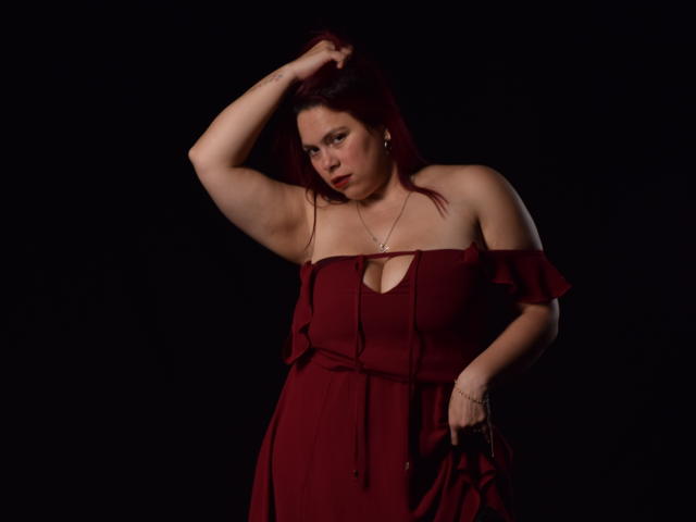 SalmaBrown with red hair, fair skin, and medium tits, wearing a red off-shoulder dress, holding her hair, looking down, dark background, slight smile, hand on hip.