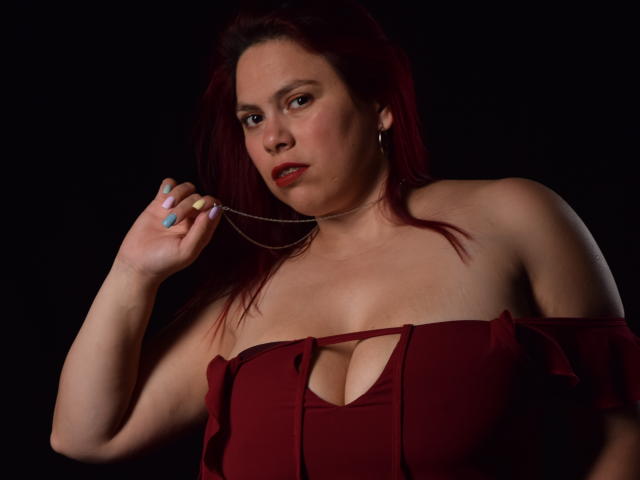 SalmaBrown with red hair, fair skin, and red lipstick, holding a chain, wearing a maroon dress with cleavage, looking at camera, dark background, visible nipples, painted nails.