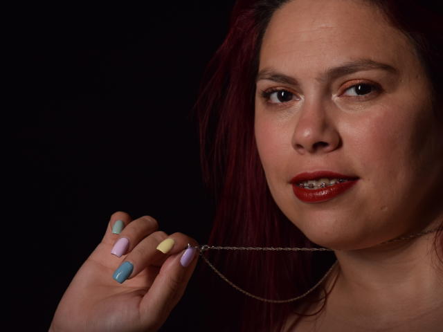 SalmaBrown con capelli rossi, rossetto rosso, che tiene una catena con unghie dipinte (rosa, giallo, blu, viola), sfondo scuro, bocca leggermente aperta, che mostra i denti, pelle chiara, occhi scuri..