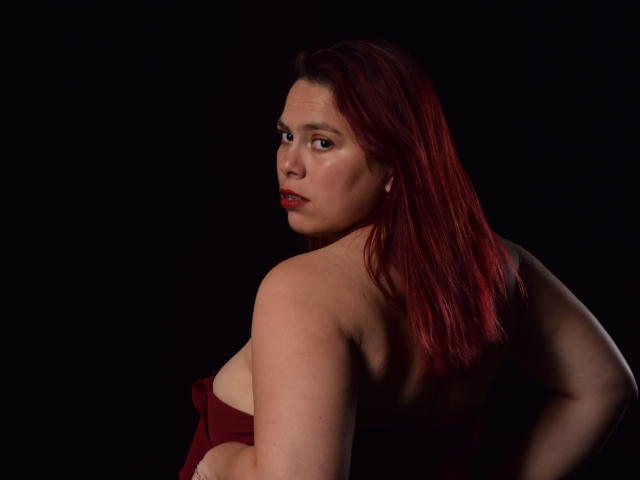 Salma Brown mit roten Haaren, die über ihre Schulter schaut und dabei ihren nackten Rücken und ihre oberen Brüste zeigt, trägt einen roten Träger und steht vor einem schwarzen Hintergrund. Sie ist leicht gedreht und hat einen ernsten Blick..