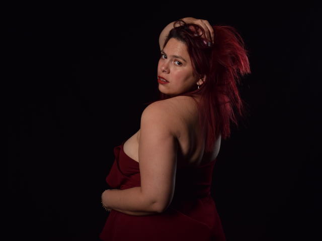 SalmaBrown with red hair, looking over shoulder, wearing a red dress, showing her back and a hint of her tits, against a black background.