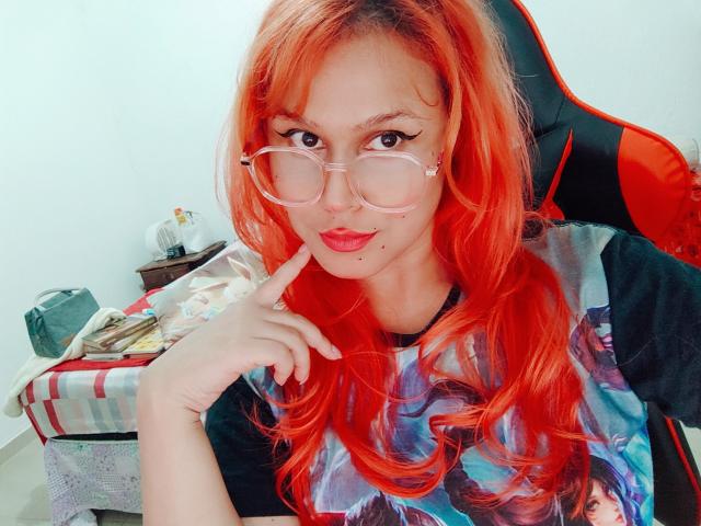 AhriValvet with bright red hair, large glasses, and a black shirt with a colorful print, touching her lip with a finger, sitting in a red and black gaming chair.