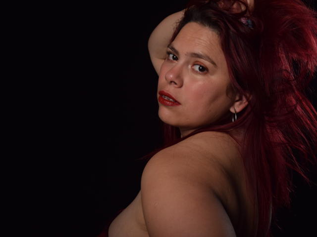 SalmaBrown with red hair, looking back over her shoulder, topless, arm raised, showing her bare tits, dark background, red lipstick, slight smile, ear hoop, dark skin.