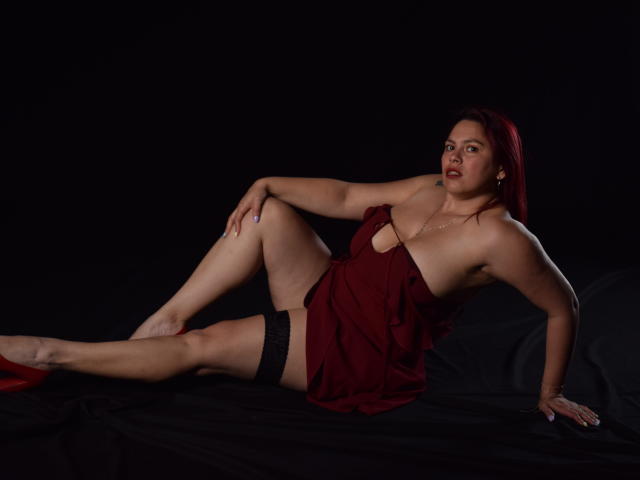 SalmaBrown è sdraiata su un panno nero, indossa un vestito rosso con un taglio, calze nere in pizzo fino alla coscia e tacchi alti rossi, con i seni e i glutei parzialmente visibili..