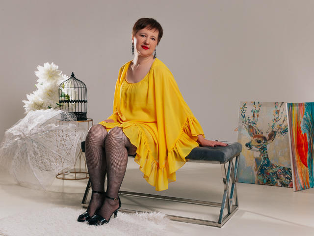 ChristaRose with short brown hair, red lipstick, yellow dress, fishnet stockings, black heels, sitting on gray bench, birdcage, white flowers, and colorful deer painting in background.