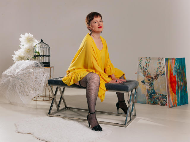 ChristaRose with short brown hair, red lipstick, yellow dress, fishnet stockings, black heels, sitting on a metal bench, yellow scarf, white flowers, birdcage, and colorful paintings.