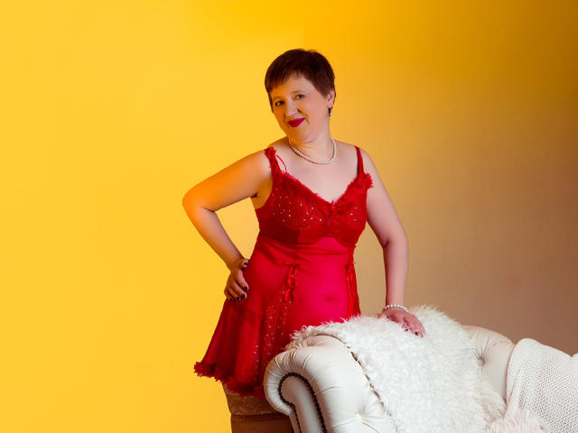 ChristaRose, short brown hair, red dress with sparkles, standing with hand on hip, white chair with fluffy blanket, yellow light, red lipstick, pearl necklace, confident pose.