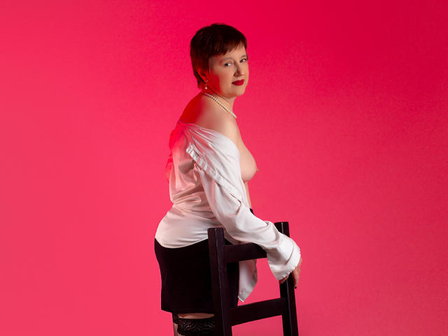 ChristaRose with short brown hair, white off-shoulder shirt, black skirt, and pearl necklace, sitting on a black chair, showing her bare tits, against a pink background.