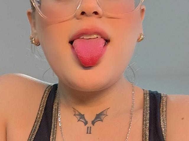 Le visage de KimSummer, avec des lunettes, la langue tirée, montrant sa langue rose. Elle a un tatouage en forme d'ailes de chauve-souris sous son cou, et elle porte un débardeur noir et des boucles d'oreilles dorées..
