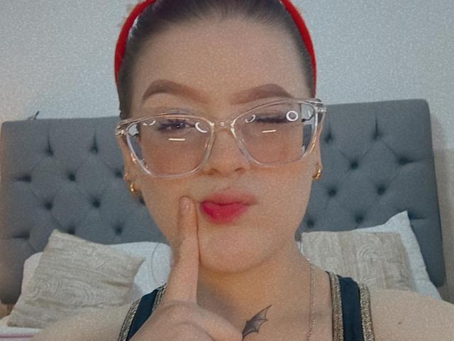 KimSummer ha una fascia rossa, occhiali trasparenti e sta indicando le sue labbra rosa e piene con un dito. Ha un tatuaggio a forma di pipistrello sul petto..