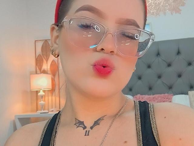 KimSummer has pink pout, clear glasses, red headband, and bat tattoo on neck, wearing gold earrings and black tank top, with a silver necklace, looking cute.