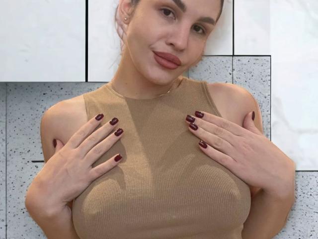 AurikaBriz with tan ribbed top, hands on her boobs, dark red nails, light skin, subtle smile, brown hair in a bun, white cabinet in background, cute.