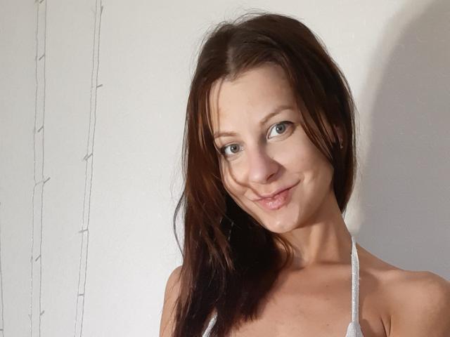 Karolina, met bruin haar, blauwe ogen en een lichte glimlach, draagt een witte top met halter, waardoor haar blote schouders en borst zichtbaar zijn. Haar borsten zijn klein en schattig..