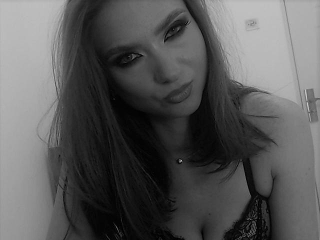 Black-and-white selfie of EllyMuller with long hair, eyeliner, and a slight pout, wearing a lace bra, showing her tits and a necklace with a small pendant.