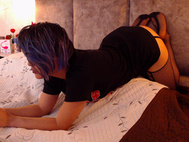 CassieJones is on a bed, on all fours, wearing a black shirt and fishnet stockings, with her ass up and purple-blue hair. Her pussy is not visible.