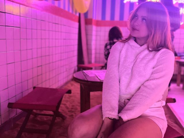 ShantalSummer with pinkish-purple hair, wearing a fluffy white bathrobe, sitting on a red bench, showing her thighs, in a neon-lit tiled room, looking to the side.