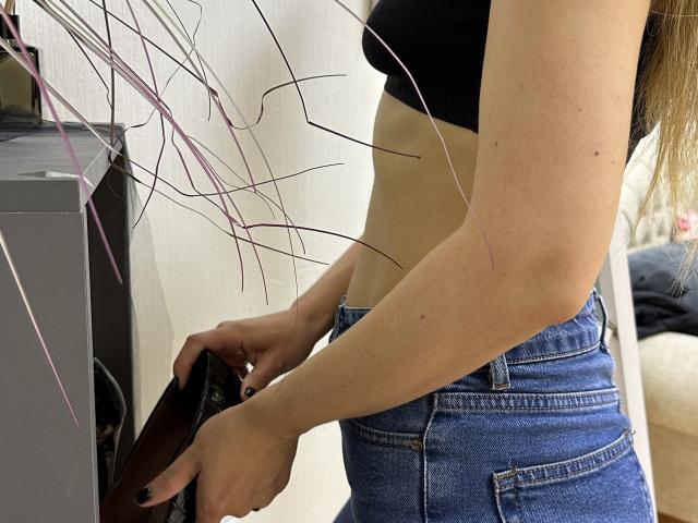 Stefanni with a slim, pale, toned midriff, wearing a black bra and blue jeans, holding a brown leather wallet, arms slightly raised, long brown hair, white wall background.