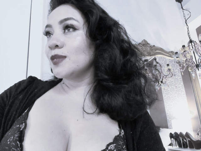 AlisGourmande with wavy black hair, wearing a black lace bra and cardigan, showing her large tits and full lips, looking to the side with a confident expression.