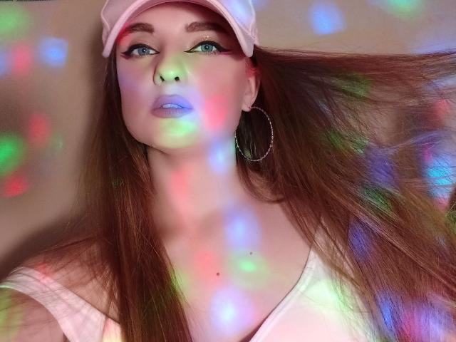 KariMillie hat langes, braunes Haar, blaue Augen und rosafarbene Lippen mit farbenfrohen Lichtreflexionen im Gesicht. Sie trägt eine rosa Mütze, Ohrringe im Hula-Hoop-Stil und ein weißes Oberteil..