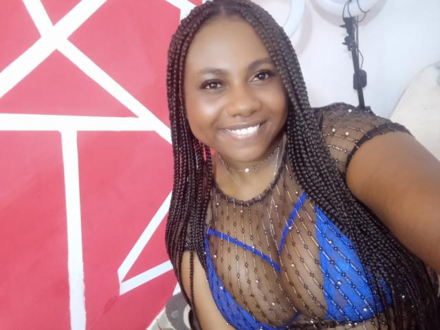 LeylaAniston with dark skin and long braided hair, smiling, wearing a sheer black top with blue mesh and sparkles, showing her tits. She has shiny white teeth.