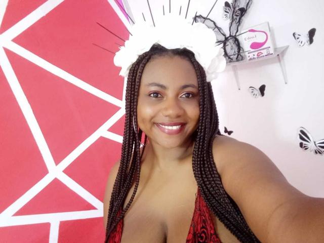 LeylaAniston with brown braided hair, white headpiece, red and black patterned top, smiling, showing her tits, medium brown skin, white background with butterfly stickers, and a pink wall.