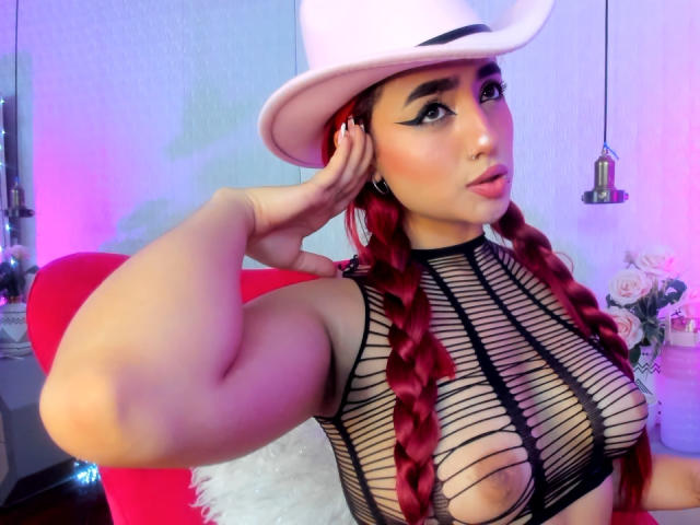 MelanieKane with red braids, wearing a white cowboy hat, black mesh top showing her tits, flexing her arm, and pouting with a nose ring.