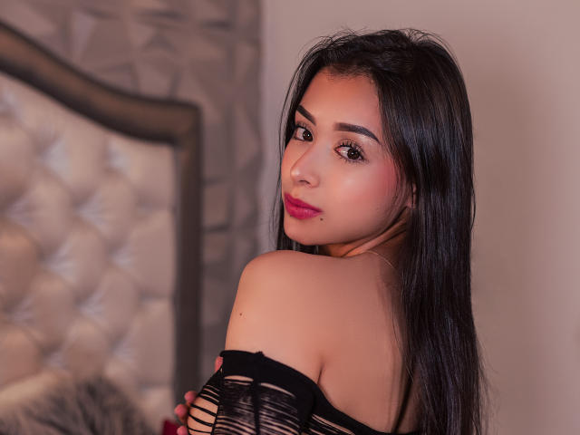 Antonella Rossy, con cabello negro largo, lápiz labial rosa y una blusa negra con un hombro descubierto, mirando por encima del hombro, mostrando sus hombros desnudos y sus pequeños senos. Iluminación suave y cálida..