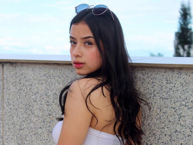 Antonella Rossy, con lunghi capelli neri, un top bianco a tubino, occhiali da sole in testa, che guarda oltre la spalla, rossetto rosa, labbra leggermente serrate, pelle chiara, piccolo piercing al naso, in piedi contro un muro con una superficie strutturata..