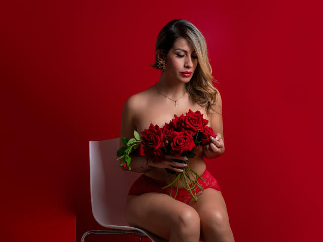 Mafeduque avec des roses rouges, portant des culottes en dentelle rouge, assise sur une chaise blanche, tenant un bouquet, sexy, fond rouge, cheveux blonds, rouge à lèvres rouge, boucles d'oreilles dorées, regardant vers le bas, mains sur les roses..