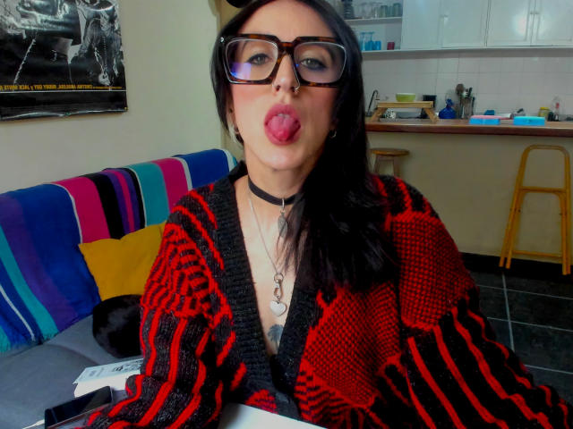 Une jeune femme aux cheveux noirs, portant des lunettes, un pull rayé rouge et noir, un collier ras du cou, la langue tirée, assise à une table, montrant ses seins, dans une cuisine, avec un canapé rayé coloré en arrière-plan..