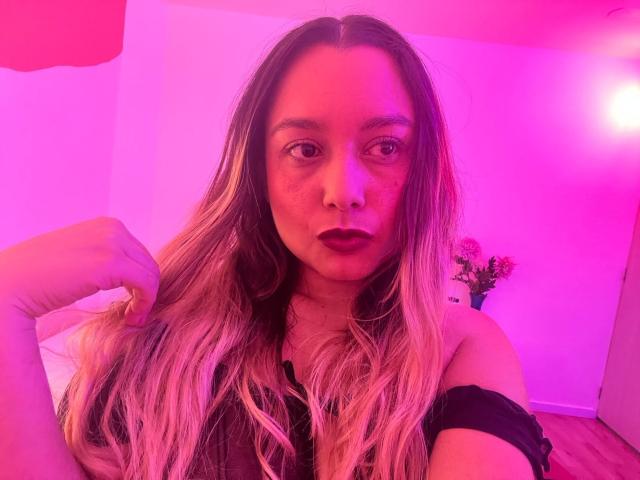 AmayaJhons met roze aanzien haar, donkere lipstick, zwarte top met een open schouder, haar vasthoudend, naar de camera kijkend, roze en rode verlichting, houten vloer, vaas met bloemen op de achtergrond..