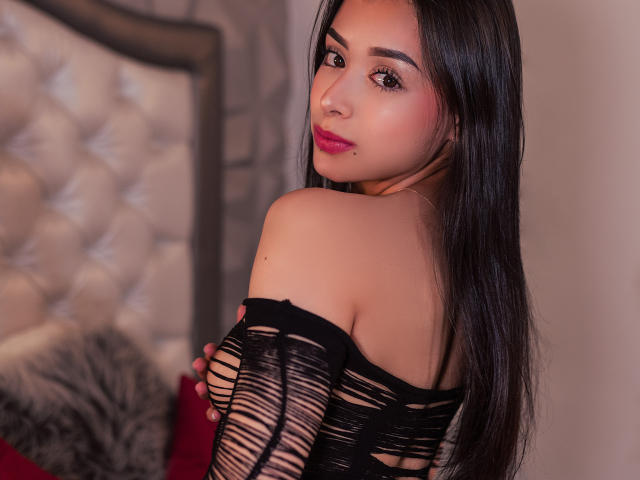 Antonella Rossy mit langen, schwarzen Haaren, trägt ein schwarzes, durchsichtiges, zerrissenes Oberteil, das ihre Schultern freilegt, und blickt mit pinkem Lippenstift und einem leichten Lächeln zurück, in einem sanft beleuchteten Schlafzimmer..