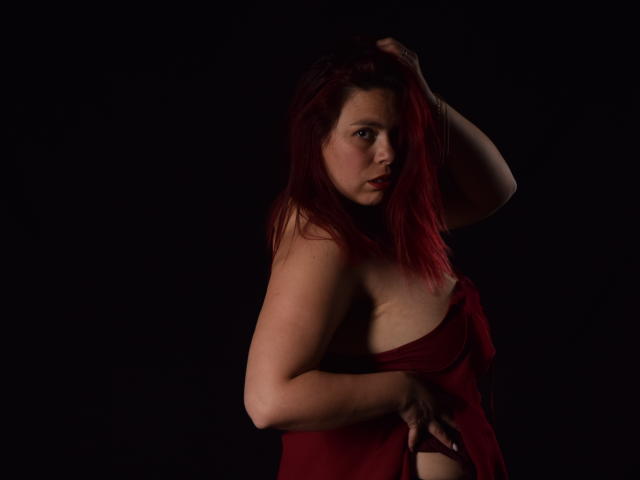 SalmaBrown, con capelli rossi, pelle chiara e seno piccolo, indossa un abito rosso avvolgente, con una mano tra i capelli, sfondo scuro, guarda la fotocamera, trucco leggero, leggero sorriso, illuminazione scura..