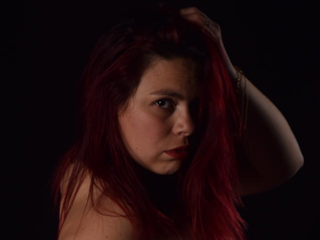 Salma Brown mit roten Haaren, die Hand im Haar, intensiver Blick, dunkler Hintergrund, dezentes Make-up, roter Lippenstift, leicht geöffneter Mund, freie Schultern, ernster Gesichtsausdruck, Armband am Handgelenk, dunkle Beleuchtung..
