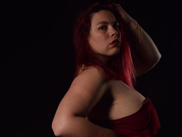 SalmaBrown, con capelli rossi, pelle chiara e seno piccolo, indossa una maglietta rossa senza spalline, con una mano tra i capelli, sfondo scuro, espressione seria, luce tenue, braccialetto dorato al polso destro..