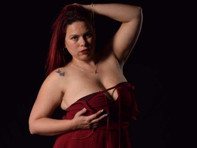 Salma Brown mit roten Haaren, trägt ein rotes, trägerloses Kleid, eine Hand am Hüfte, die andere Hand im Haar, zeigt Brüste, Tattoo an der linken Schulter, schwarzer Hintergrund..