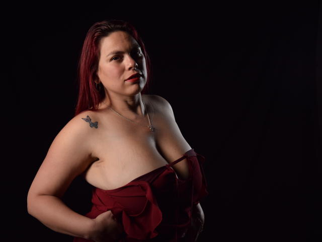SalmaBrown with red hair, pale skin, small tattoos on shoulders, wearing a strapless red dress, holding it down to show her tits, dark background, looking at camera.