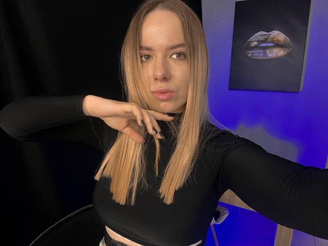 CandyWithBrandy, com cabelos loiros longos, blusa preta de manga comprida e lábios carnudos, apontando para a câmera, exibindo seus pequenos seios, em um selfie com iluminação azul..