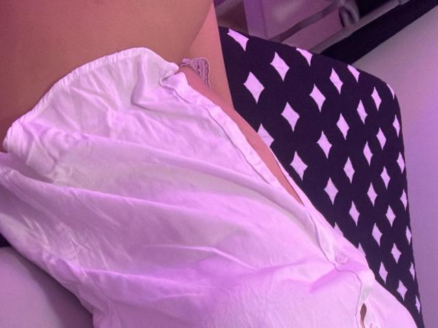 AmayaJhons' white shirt is pulled up, exposing her bare ass and a glimpse of her white lace panties. The shirt has a purple tint, and she's on a black and white patterned seat.