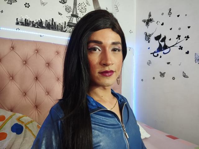 MilyPower, avec de longs cheveux noirs, portant une chemise bleue à fermeture éclair, un tête de lit rose avec des motifs en relief, et du rouge à lèvres violet, dans une chambre avec des autocollants muraux de papillons et de la Tour Eiffel..