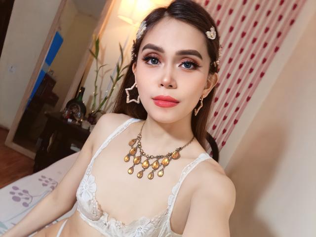 TSAnabelleSwift with dark hair, wearing a white lace bra, gold necklace, and star-shaped earrings, looking at the camera with red lipstick and blue eyes. Boobs visible.
