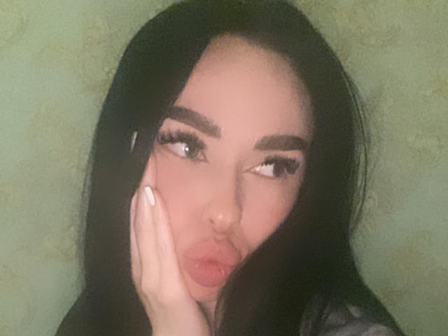 BellaMegan, avec de longs cheveux noirs, les lèvres pleines légèrement pincées, une main couvrant la moitié de son visage, regardant sur le côté, sourcils foncés, longs cils, fond vert, lumière douce, légèrement flou..