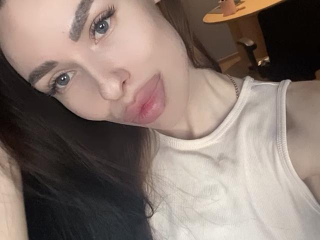 BellaMegan hat volle, rosafarbene Lippen, dicke Augenbrauen, langes, dunkles Haar und helle Haut. Sie trägt ein weißes Top ohne Ärmel, das ihre nackten Schultern zeigt. Ihre blauen Augen sind leicht geöffnet..