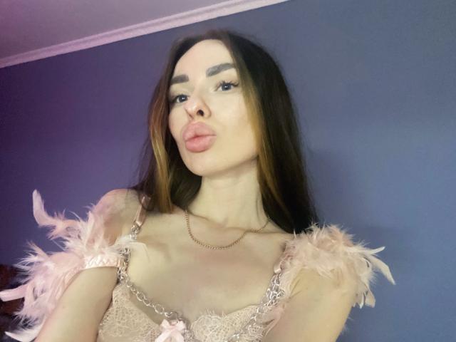 BellaMegan, con cabello castaño largo, labios rosados y sensuales, vestida con una blusa rosa cubierta de plumas, tiene senos pequeños, piel clara y un collar de cadena de oro, mirando a la cámara..