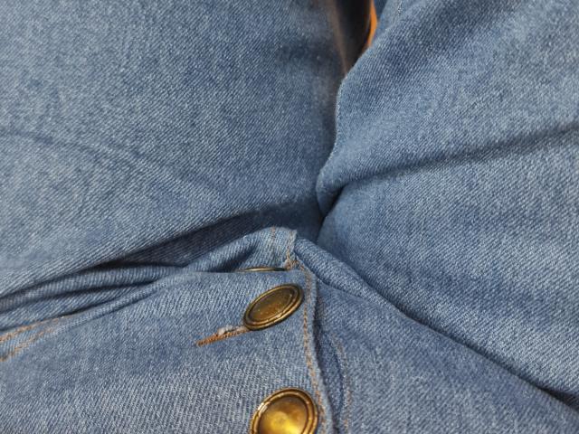 De blauwe spijkerbroekbillen van MonaCool, met zichtbare stoftextuur en twee koperen knopen, licht gekreukeld, gecentreerd in het beeld..