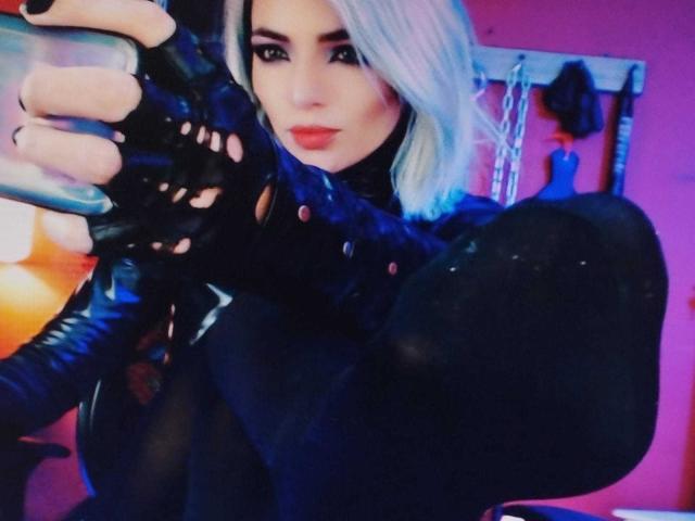 MisssDarkLady with platinum blonde hair, wearing black latex and fingerless gloves, holding a dildo, her large black-covered boot in the foreground, pink walls in the background.