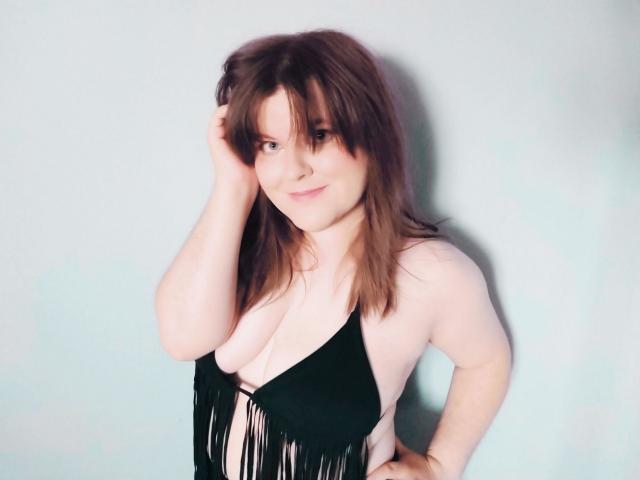 Dahlia, cabelo castanho, mão no cabelo, vestindo blusa preta com franjas, mostrando os seios, pele clara, sorriso discreto, em pé contra um fundo claro..