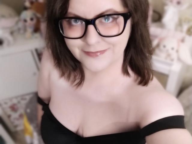 DahliaGray, portant des lunettes à monture noire, sourit à l'appareil photo. Elle porte un haut noir à une épaule, laissant entrevoir sa poitrine, et a les cheveux bruns mi-longs. Elle se trouve dans une pièce éclairée par une lumière douce..